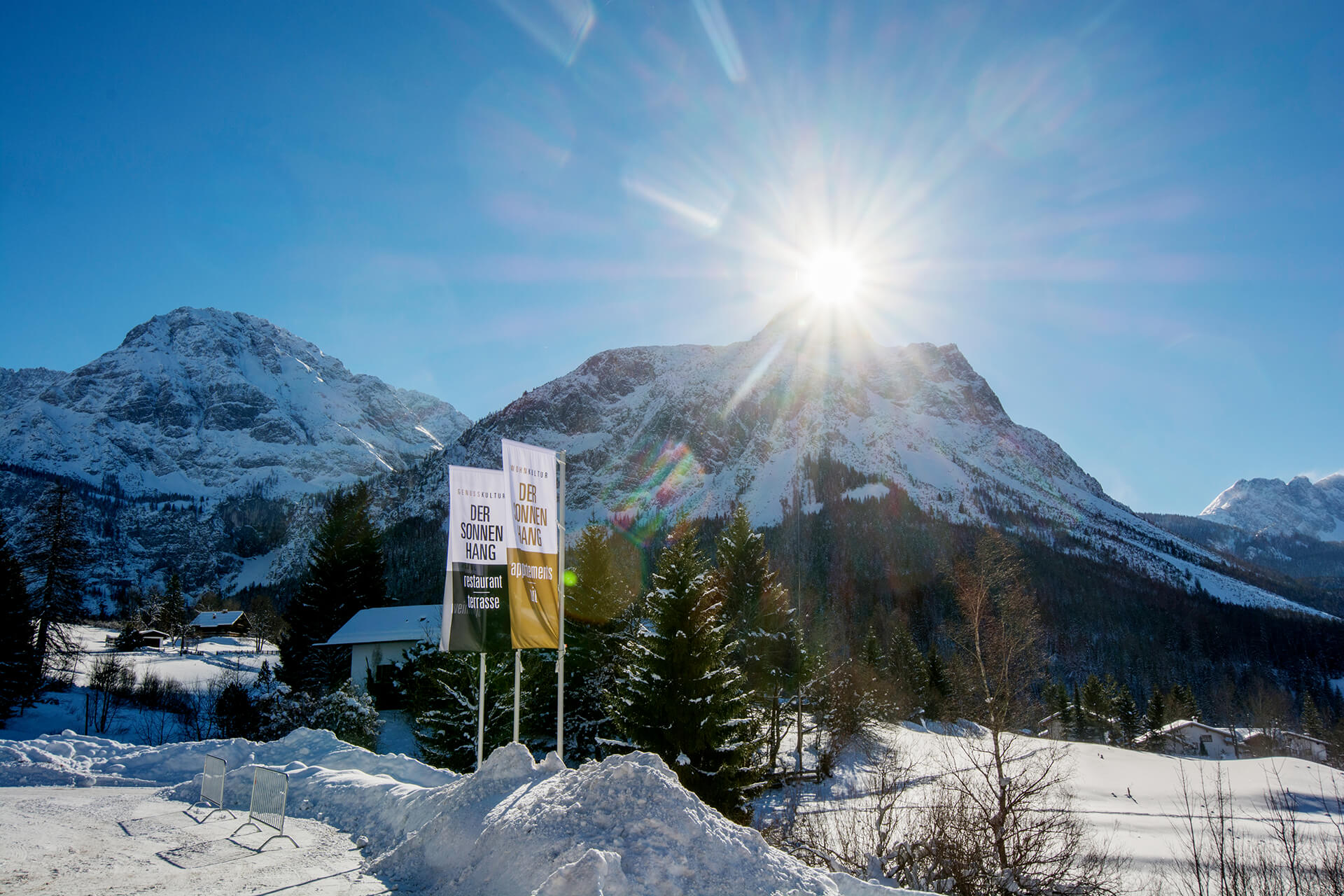 Blick auf die Berge, Schneehaufen am Parkplatz vor der Terrasse. Sonnenschein über dem Berggipfel.